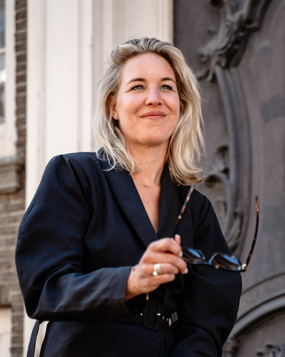 Robin Visser-Ardon in een donkere blazerjurk, zittend op een smeedijzeren hek met haar zonnebril in de hand
